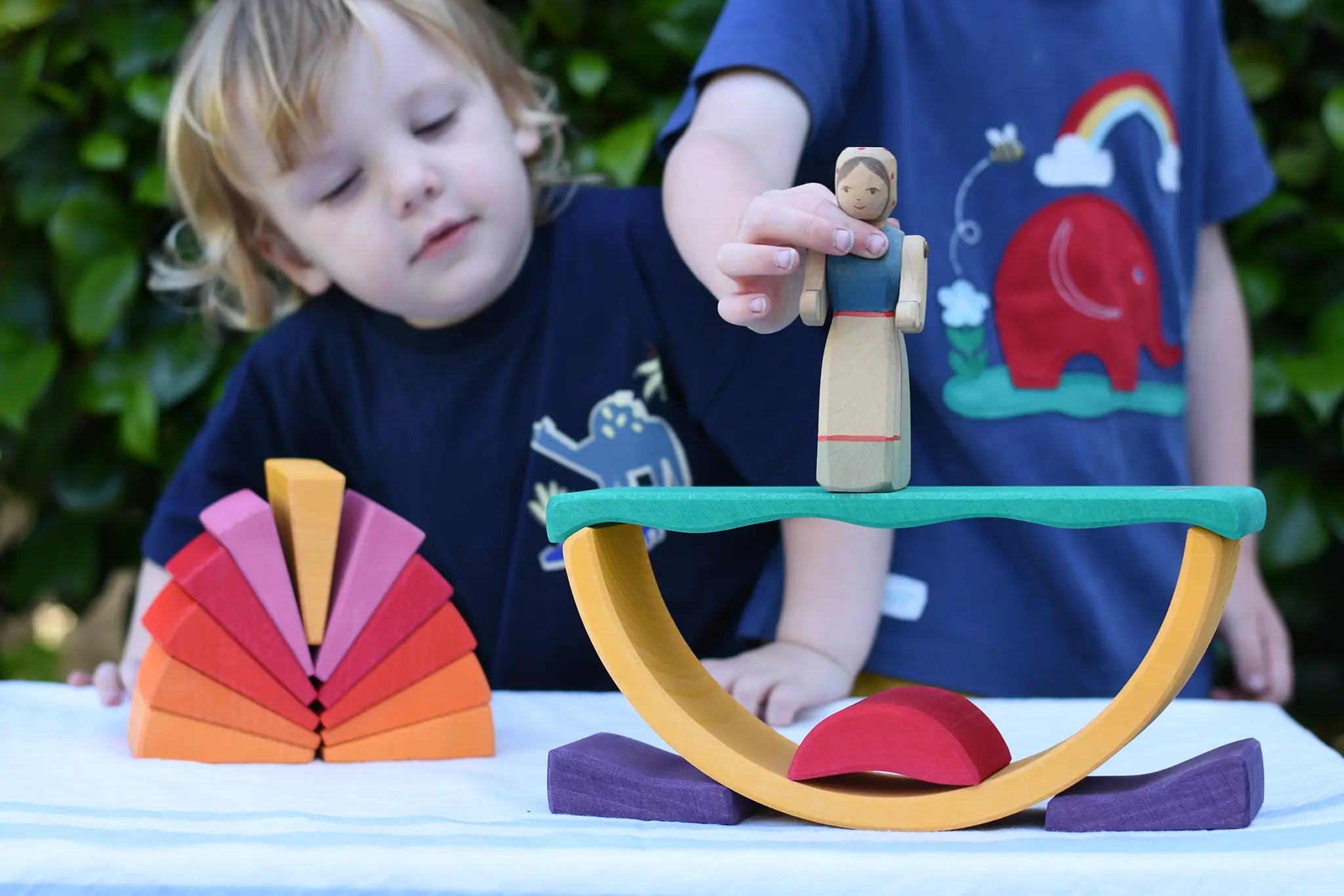 Children playing with the Glückskäfer wooden stacking sunray arch in yellow purple placed on a table outdoors. They are placing an Ostheimer wooden figure on top.