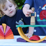 Children playing with the Glückskäfer wooden stacking sunray arch in yellow purple placed on a table outdoors. They are placing an Ostheimer wooden figure on top.