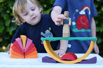 Children playing with the Glückskäfer wooden stacking sunray arch in yellow purple placed on a table outdoors. They are placing an Ostheimer wooden figure on top.