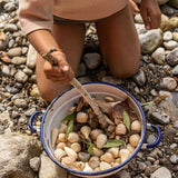 A child playing with the Grapat 120 Natural Wooden Mandala Pieces, placing them into a metal bowl with leaves and mixing with a wooden stick. A mix of 10 different shapes in a natural wood. There are 12 of each shape supplied. These wooden loose part toys are part of a wide range of open ended toys available at Babipur. 