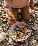 A child playing with the Grapat 120 Natural Wooden Mandala Pieces, placing them into a metal bowl with leaves and mixing with a wooden stick. A mix of 10 different shapes in a natural wood. There are 12 of each shape supplied. These wooden loose part toys are part of a wide range of open ended toys available at Babipur. 
