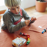 Child's threading colourful wooden beads onto a string from one of the gifts included in the Grapat 2026 advent calendar available at Babipur. The colourful gift box can be seen in front of the child.