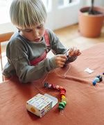Child's threading colourful wooden beads onto a string from one of the gifts included in the Grapat 2026 advent calendar available at Babipur. The colourful gift box can be seen in front of the child.