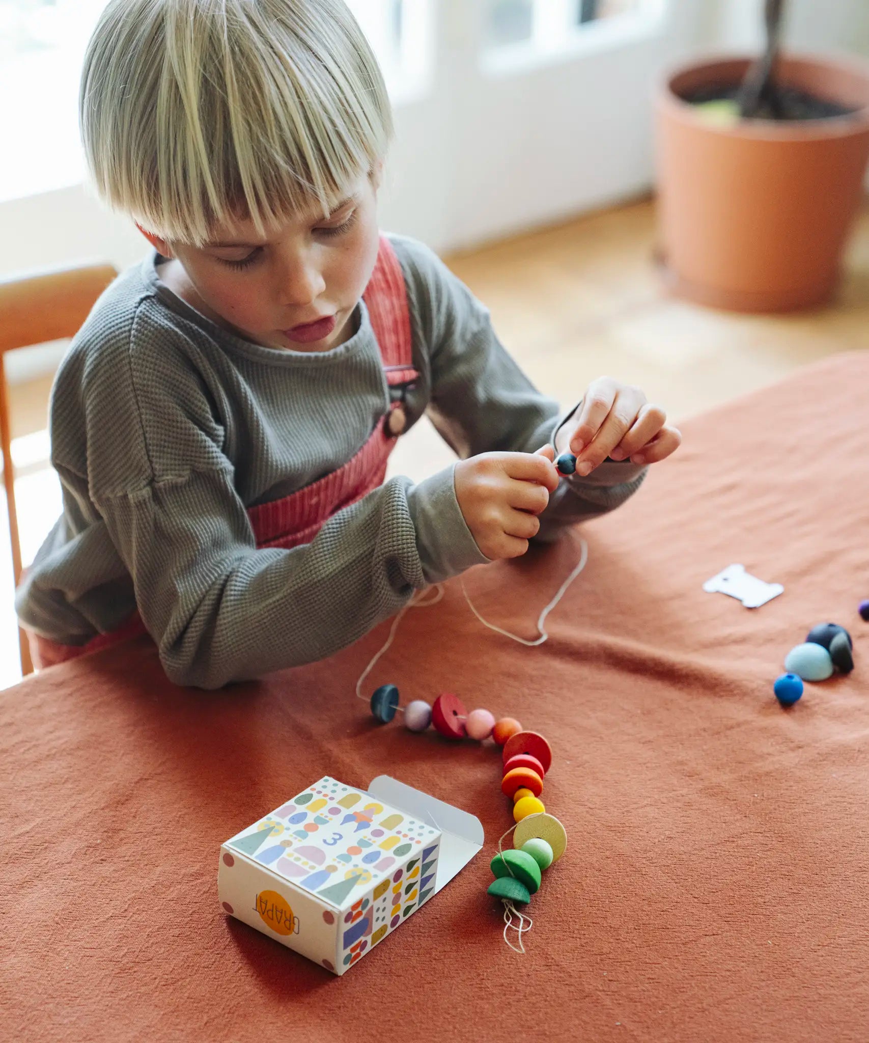 Child's threading colourful wooden beads onto a string from one of the gifts included in the Grapat 2026 advent calendar available at Babipur. The colourful gift box can be seen in front of the child.