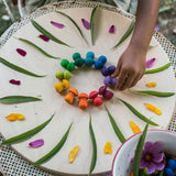 A close up of a child's hand placing mushrooms from the Grapat 36x Mandala Rainbow Mushrooms set in a circular shape to create a mandala. These wooden toys are part of a wide range of open ended toys available at Babipur.  