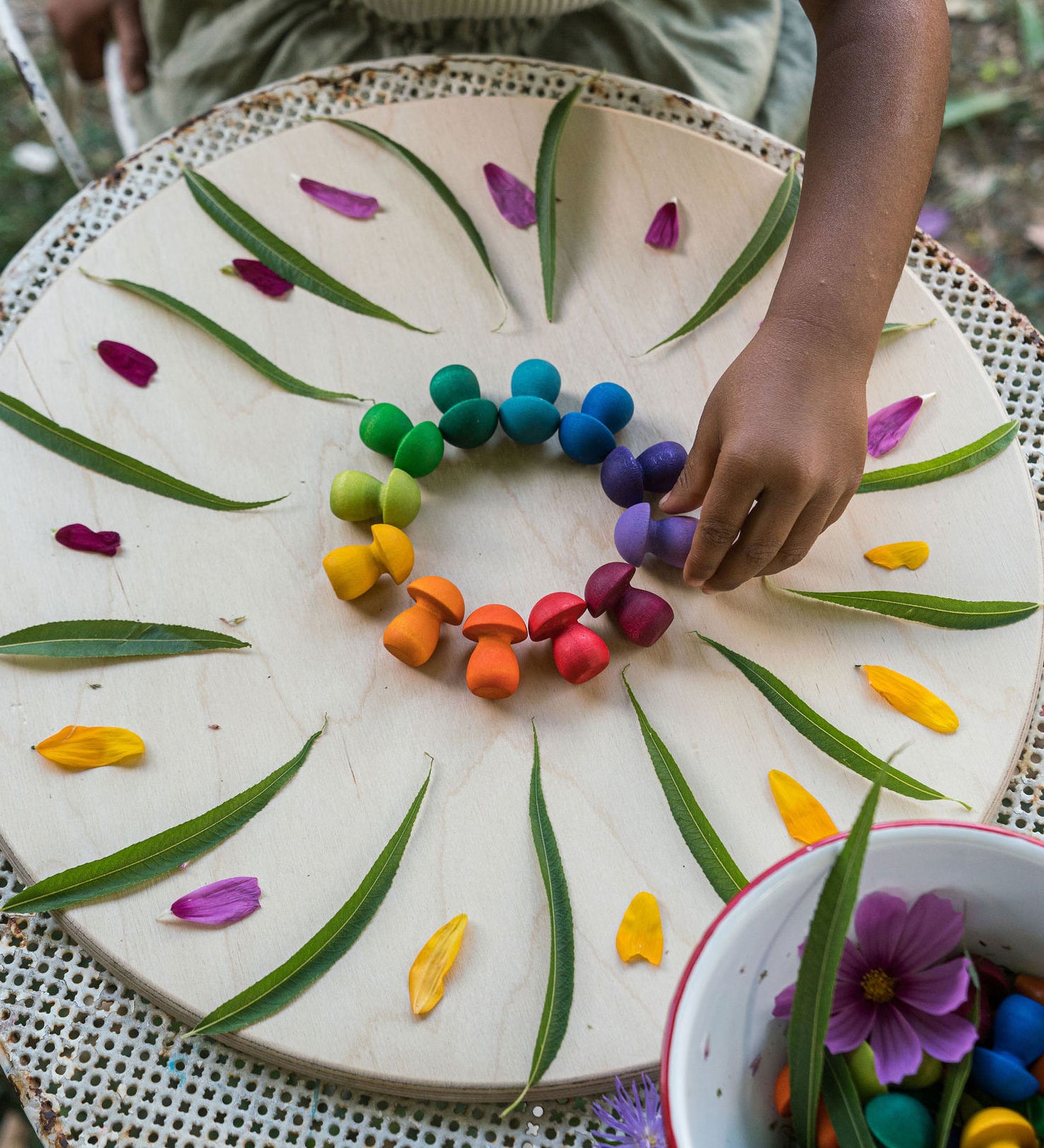 A close up of a child's hand placing mushrooms from the Grapat 36x Mandala Rainbow Mushrooms set in a circular shape to create a mandala. These wooden toys are part of a wide range of open ended toys available at Babipur.  