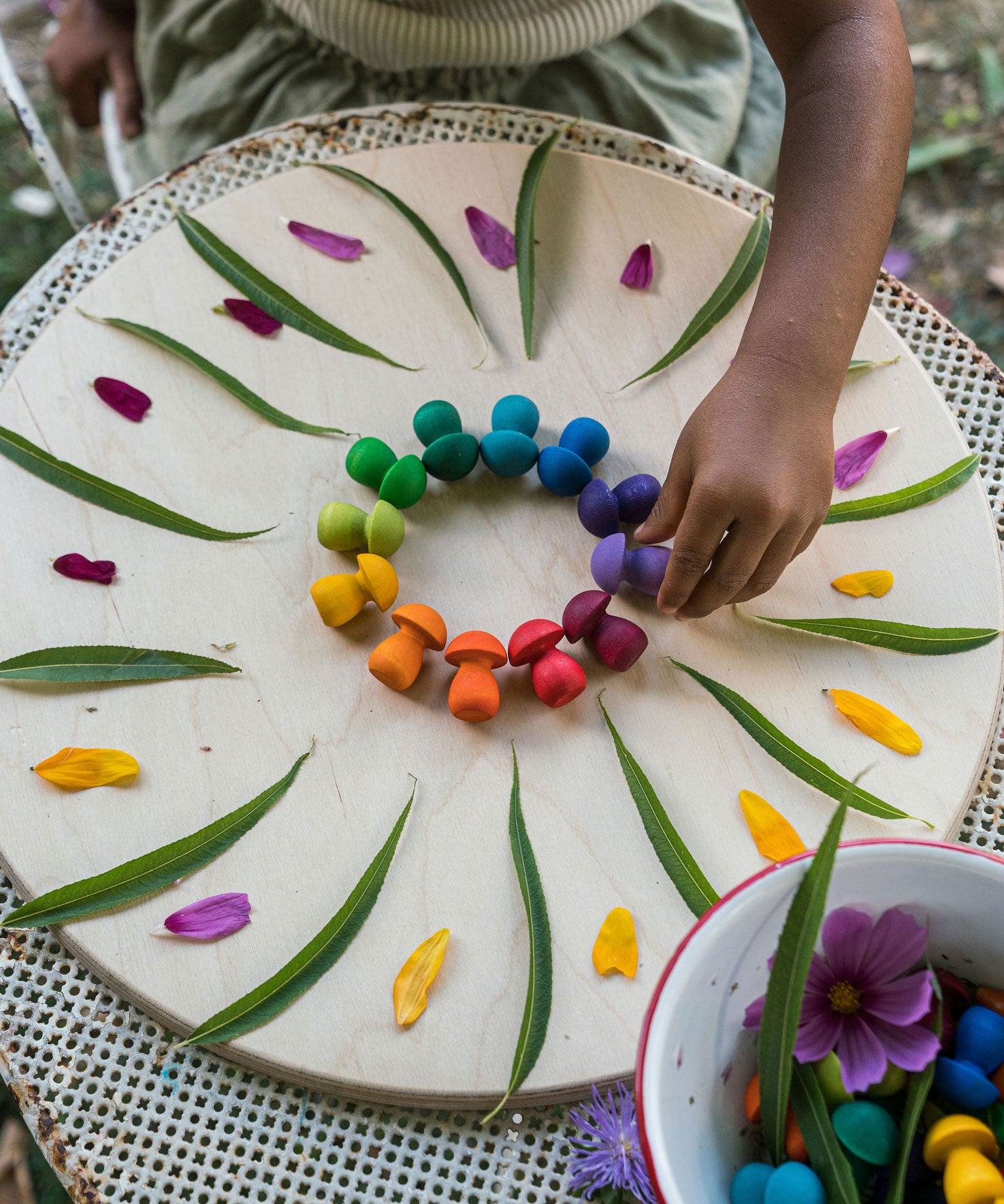 A close up of a child's hand placing mushrooms from the Grapat 36x Mandala Rainbow Mushrooms set in a circular shape to create a mandala. These wooden toys are part of a wide range of open ended toys available at Babipur.  