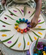 A close up of a child's hand placing mushrooms from the Grapat 36x Mandala Rainbow Mushrooms set in a circular shape to create a mandala. These wooden toys are part of a wide range of open ended toys available at Babipur.  