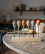 Grapat wooden twin soul puppets on a kitchen table resting on a wooden stand showing the different handcrafted faced puppets. Features a grey, brown, orange, blue handcrafted wooden puppet. 