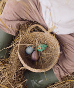 The Grapat Handmade Wooden Aurora Eggs Cold Colours Easter Decorations placed in a jute handled basket with bits of straw inside and around it. . 