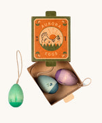 Grapat Handmade Wooden Aurora Eggs Cold Colours Easter Decorations. The eggs are pictures alongside the cardboard box packaging which has a picture of a gen on the front. A grren coloured egg is shown out of the box with the purple and blue eggs inside the box.