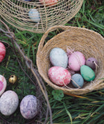 The Grapat Handmade Wooden Aurora Eggs Cold Colours Easter Decorations placed in a jute basket next to real dyed eggs. 