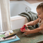 Child standing on a stone floor next to a wooden table with books and a comb on the table in the bathroom. Child playing with the Grapat wooden pink wobbly bird showing the hand painted purple feather detailing 