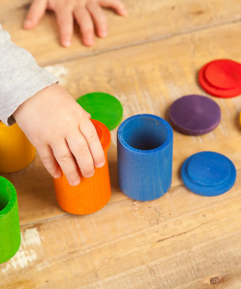 Child hand holding a orange Grapat handcrafted wooden cup with lid next to other coloured cups on a table