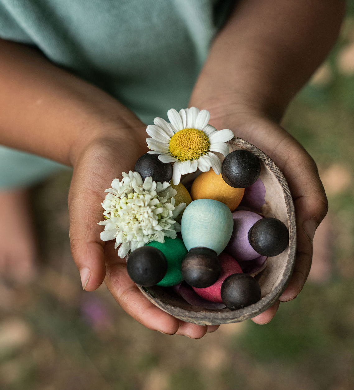 Grapat 6 Dark Baby Nins. A set of 6 small dark wooden baby Nin peg dolls in half a coconut shell, held in a child's hands. Grapat have a wide range of wooden toys and loose parts available at Babipur. 