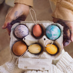 The Grapat Handmade Sunrise Eggs Wooden Easter Decorations shown placed in a cardboard egg tray next to 3 real eggs that have been painted in various colours. 