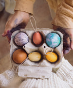The Grapat Handmade Sunrise Eggs Wooden Easter Decorations shown placed in a cardboard egg tray next to 3 real eggs that have been painted in various colours. 