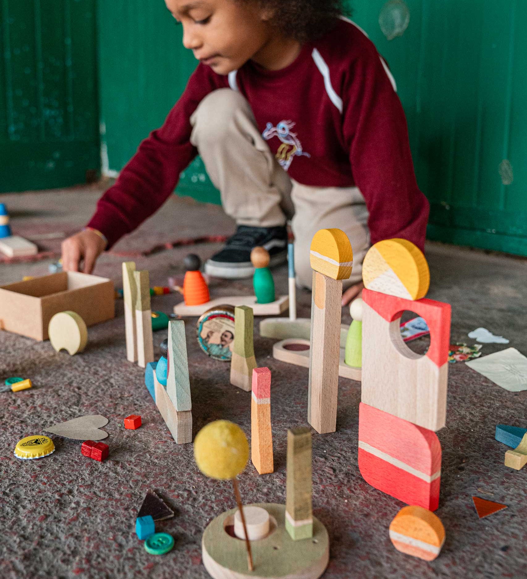 A child playing with pieces from the Grapat Happy Place Wooden Play Set. This set contains a mix of loose parts in different shapes and colours. These wooden loose part toys are part of a wide range of open ended toys available at Babipur. 