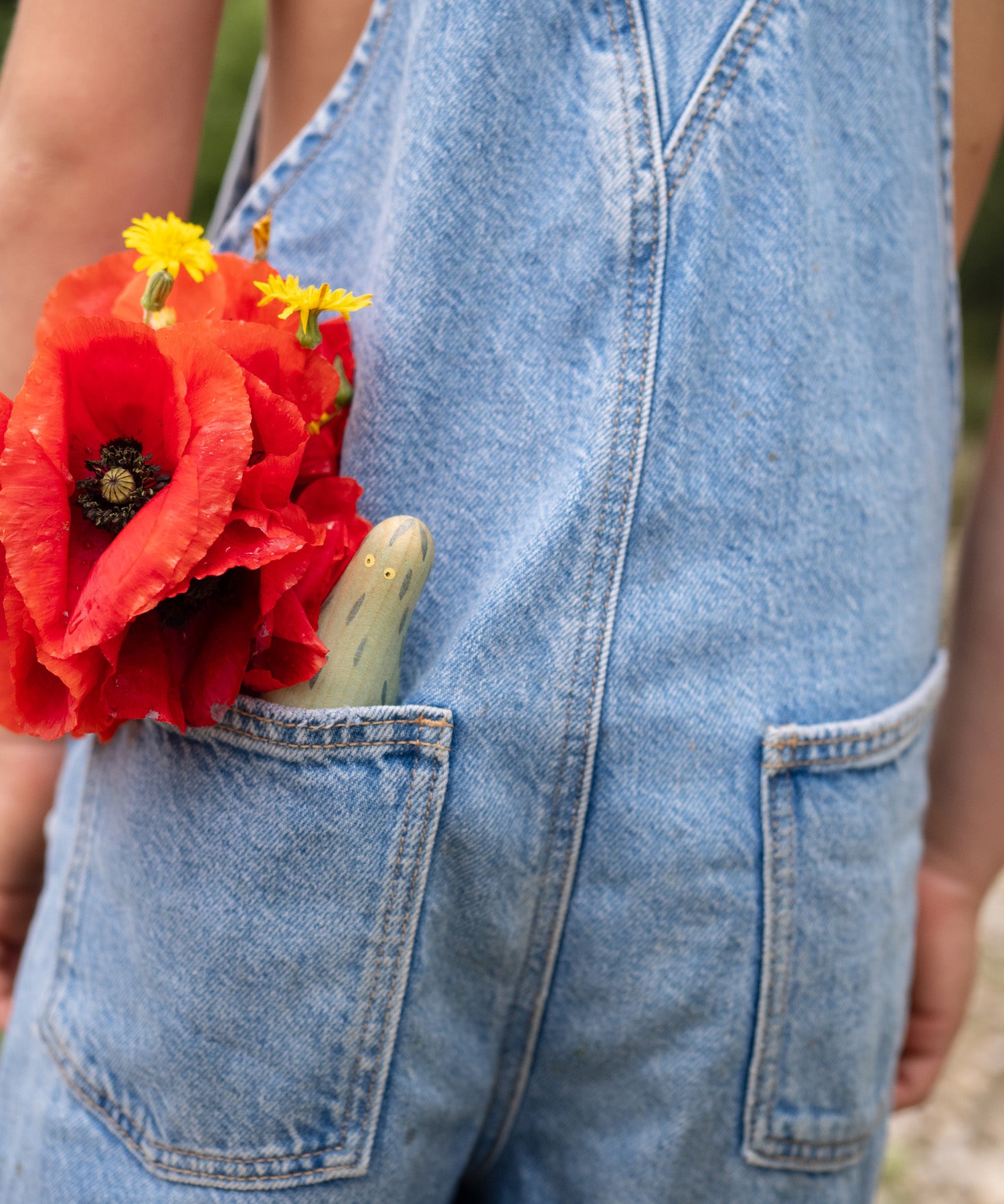 A close up of the wooden monster peg dolls from the Grapat lucky lucky box in a pocket next to a red flower showing the delicate and playful hand painted design 