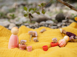 A decorative outdoor setting showing the 
 pink wooden peg dolls and pieces from the Grapat lucky lucky boxes standing on a yellow cloth. Features delicate and playful hand painted designs. 