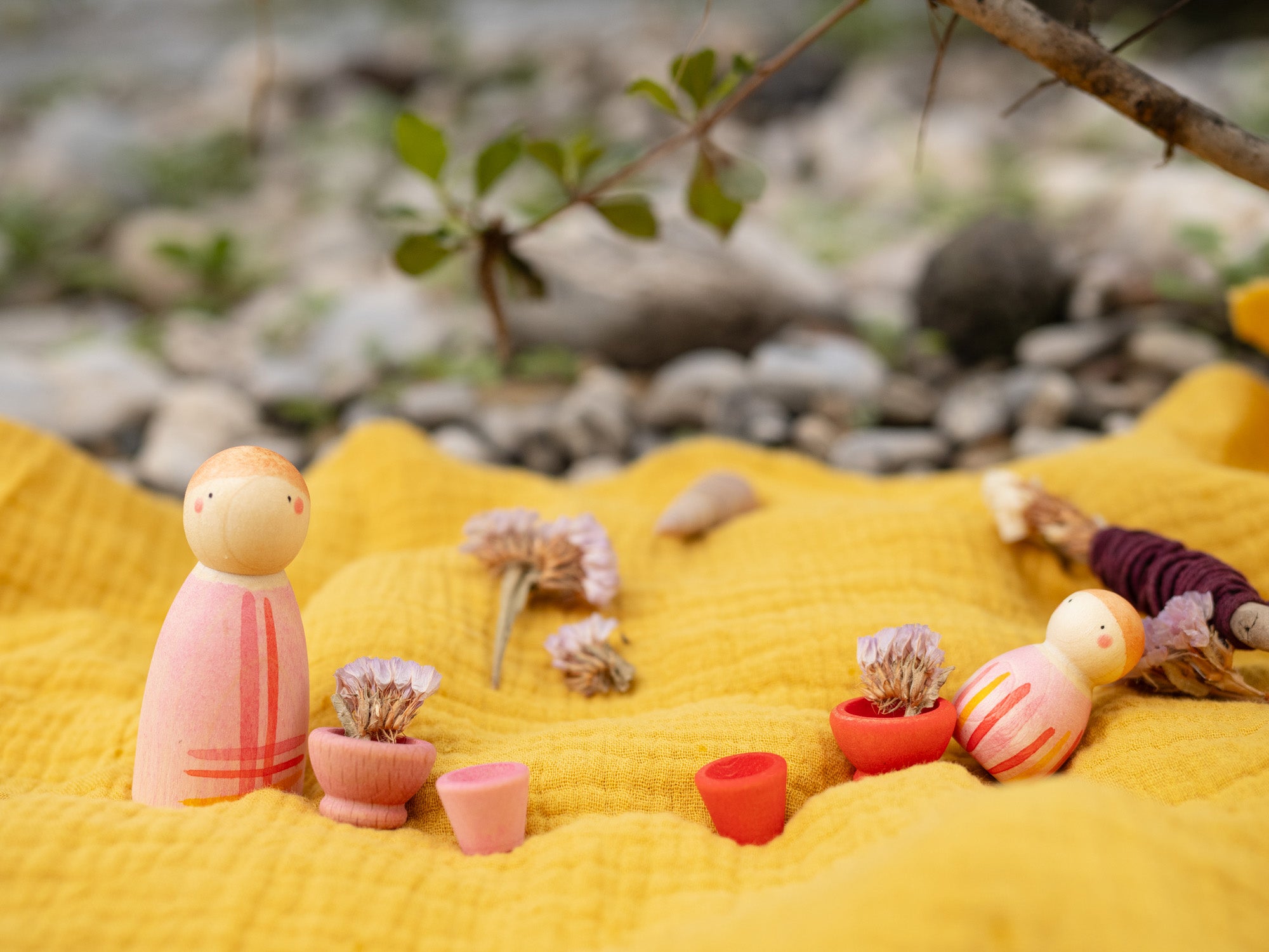 A decorative outdoor setting showing the 
 pink wooden peg dolls and pieces from the Grapat lucky lucky boxes standing on a yellow cloth. Features delicate and playful hand painted designs. 