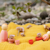 A decorative outdoor setting showing the 
 pink wooden peg dolls and pieces from the Grapat lucky lucky boxes standing on a yellow cloth. Features delicate and playful hand painted designs. 
