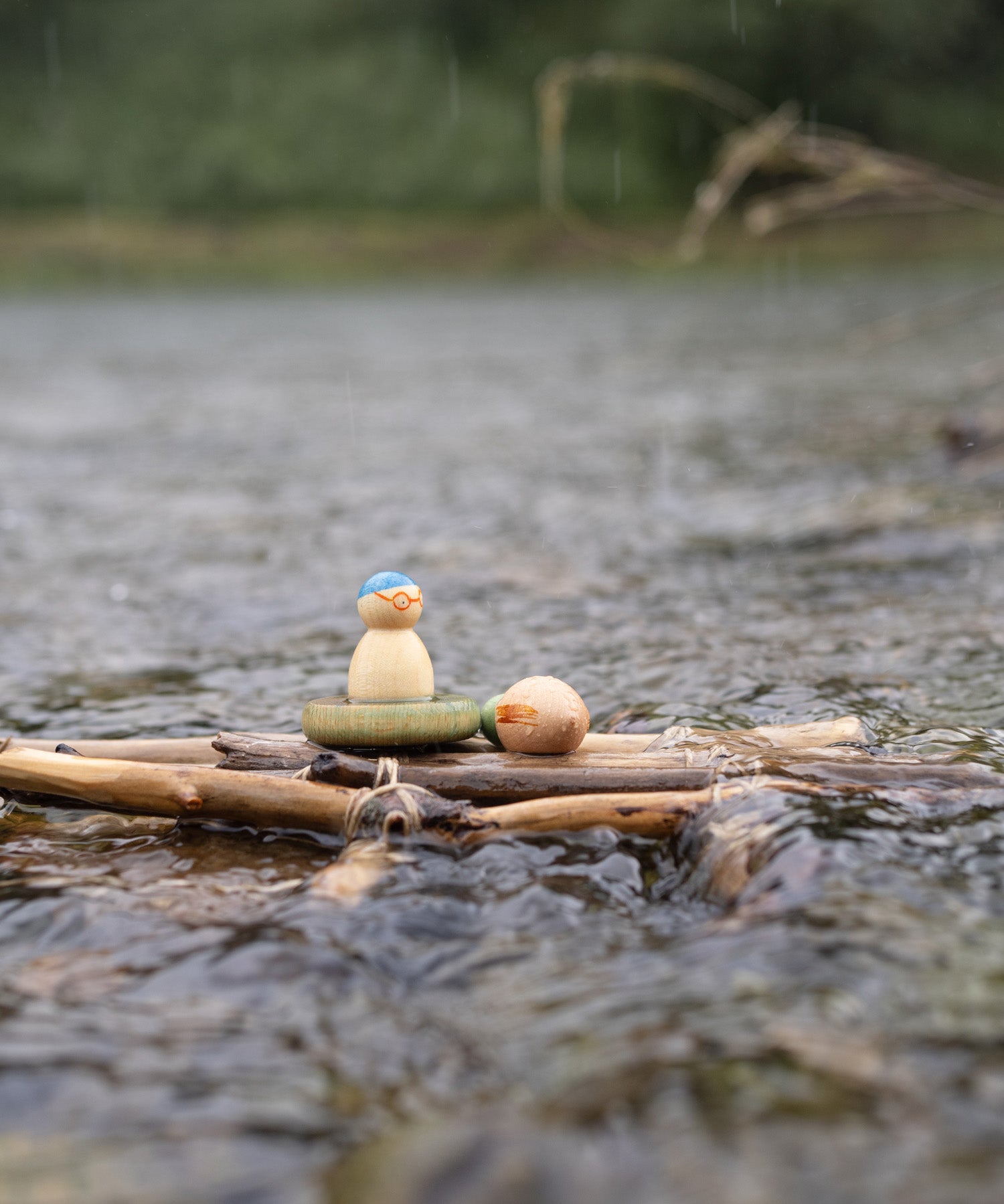 A rainy outdoor setting showing the 3 swimming inspired wooden peg dolls and pieces from the Grapat lucky lucky boxes on a bunch of sticks. 