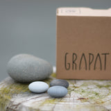 Grapat Mandala Stones placed next to a grey stone and the packaging box. A set of 36 pebble shaped wooden mandala pieces in three shades of grey, 12 in each shade. Grapat have a wide range of wooden toys and loose parts available at Babipur. 