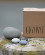 Grapat Mandala Stones placed next to a grey stone and the packaging box. A set of 36 pebble shaped wooden mandala pieces in three shades of grey, 12 in each shade. Grapat have a wide range of wooden toys and loose parts available at Babipur. 