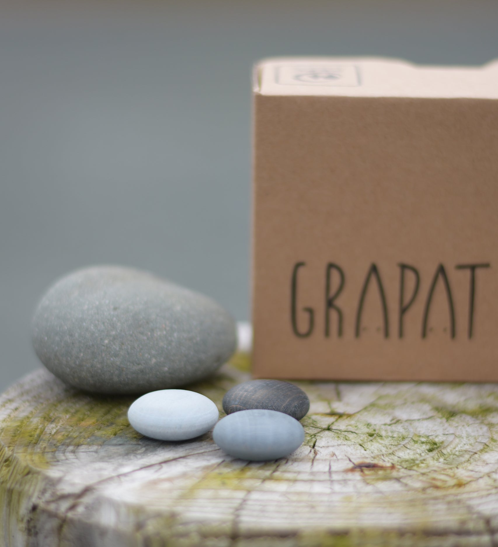 Grapat Mandala Stones placed next to a grey stone and the packaging box. A set of 36 pebble shaped wooden mandala pieces in three shades of grey, 12 in each shade. Grapat have a wide range of wooden toys and loose parts available at Babipur. 