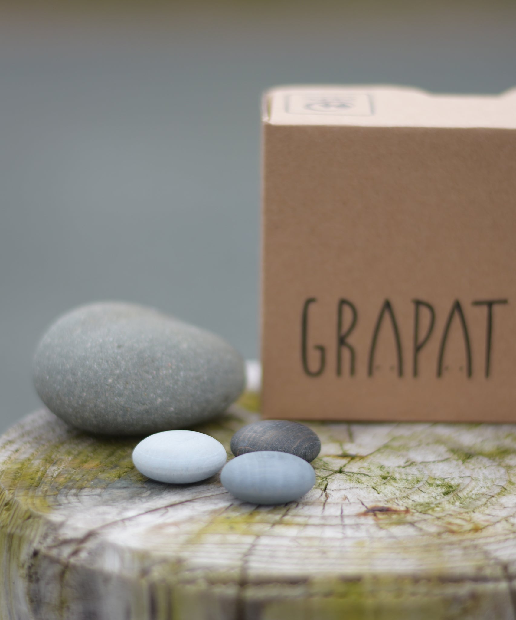Grapat Mandala Stones placed next to a grey stone and the packaging box. A set of 36 pebble shaped wooden mandala pieces in three shades of grey, 12 in each shade. Grapat have a wide range of wooden toys and loose parts available at Babipur. 