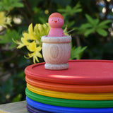 Grapat mate wooden loose part cup with animal crew peg doll placed inside placed on a stack of rainbow dishes.