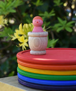 Grapat mate wooden loose part cup with animal crew peg doll placed inside placed on a stack of rainbow dishes.