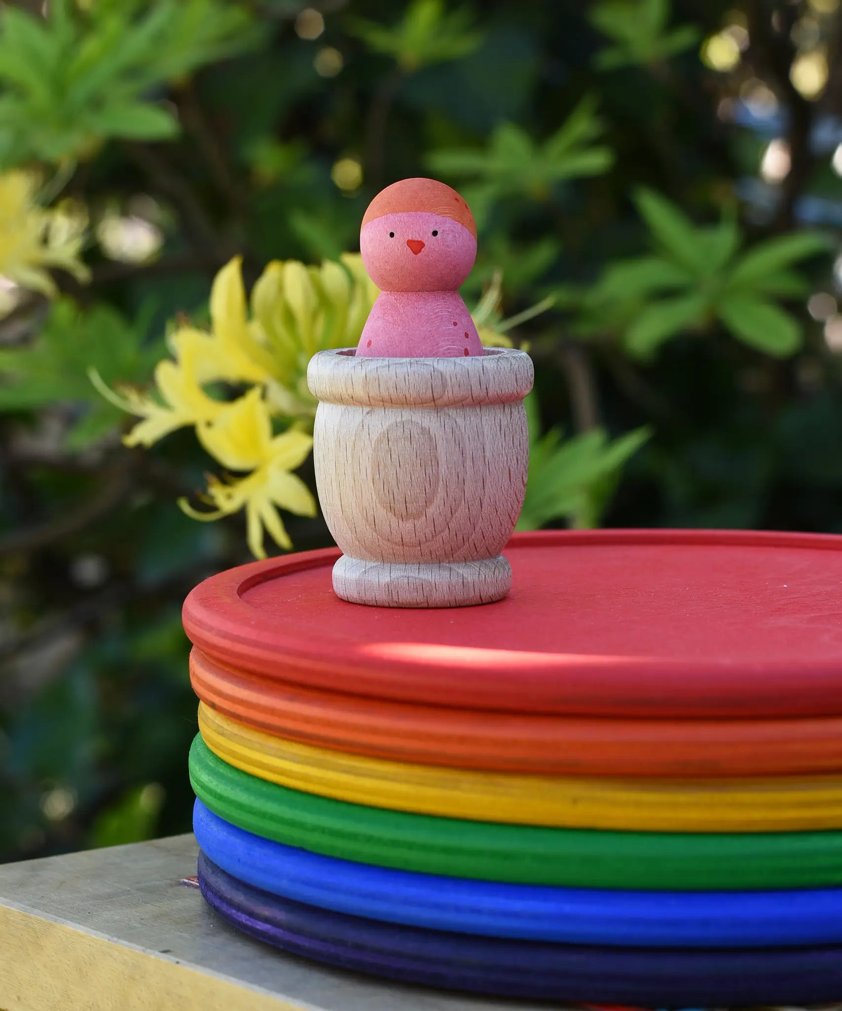 Grapat mate wooden loose part cup with animal crew peg doll placed inside placed on a stack of rainbow dishes.