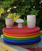 Grapat loose parts, mates, mushroom, honeycomb cup placed on a stack of rainbow dishes. 