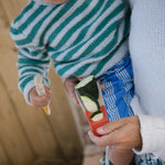 A child sorting various seeds and food items whilst holding onto one of the Grapat Wooden Sensory Play Tools. Different foods lay on the table which is covered in a blue and white chequred table cloth