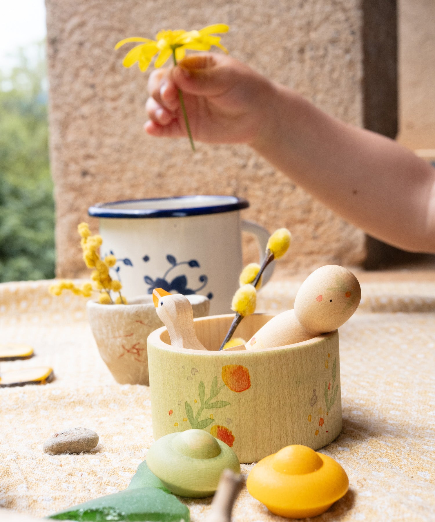 A close up of the Grapat / Ostheimer Hello Summer set showing the wooden goose and peg doll sitting inside the small doll with the 2 mandala pieces in front. Showing a child's hand holding a flower in background. 