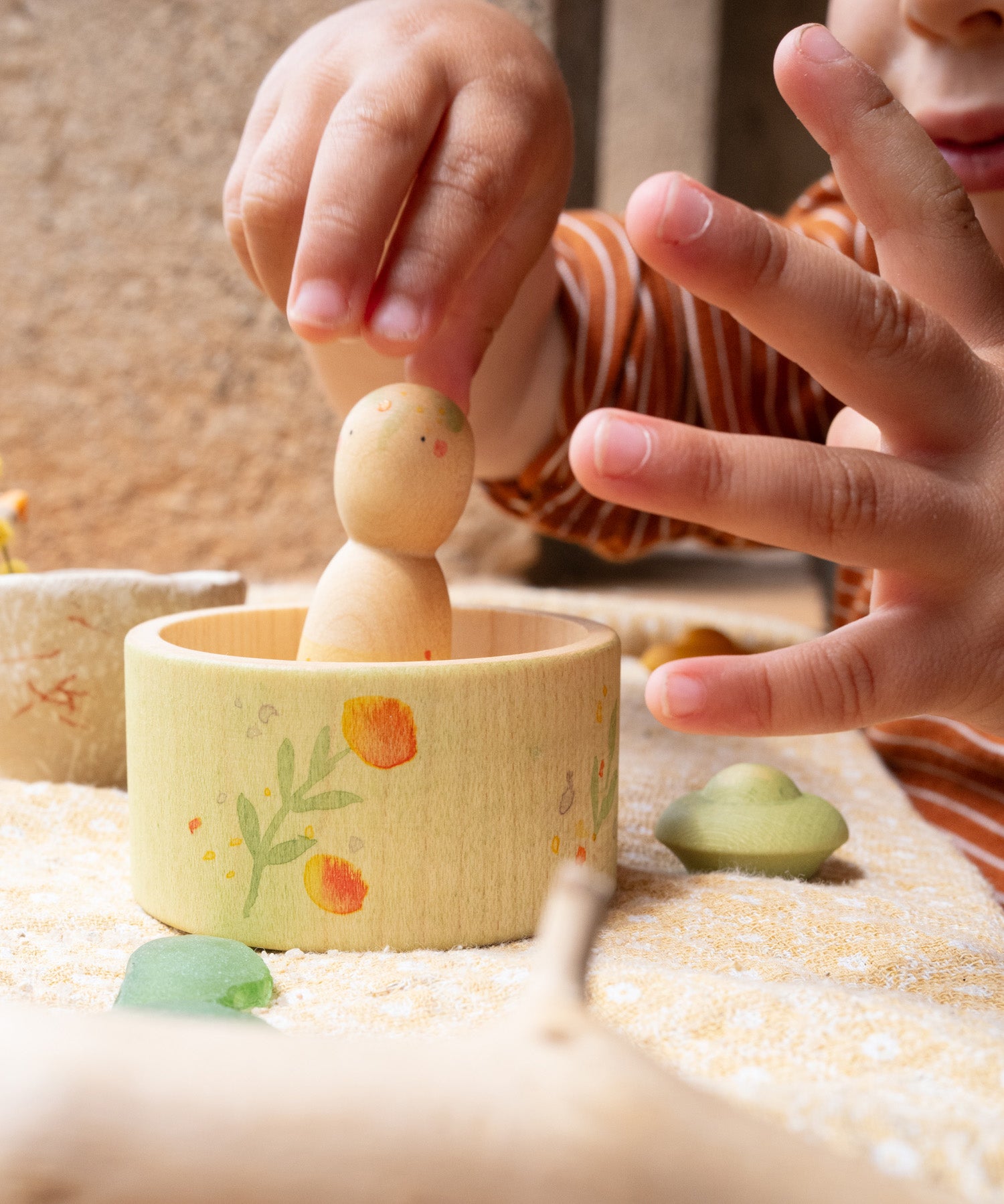 A child's hand picking up the wooden peg doll standing inside the small bowl from the Grapat / Ostheimer Hello Summer set showing the scale and delicate hand painted detail.