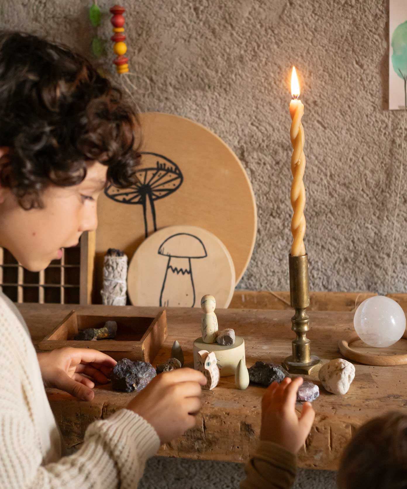 The Grapat Ostheimer Limited Edition Hello Winter wooden toy set available at Babipur. Features 5 hand cut and hand painted figures and pieces including a peg doll, small decorative bowl, owl and 2 green coloured snowdrop mandala pieces. The pieces from the set have been placed on a wooden surface and surrounded by bit of rocks and crystals. Two children are standing in front of them reaching for some of the pieces.