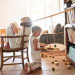 A child playing with the Grapat wooden pendulum set with wooden blocks on a terracotta floor next to a child sitting on a wooden chair with different handcrafted blocks scattered on the terracotta floor.