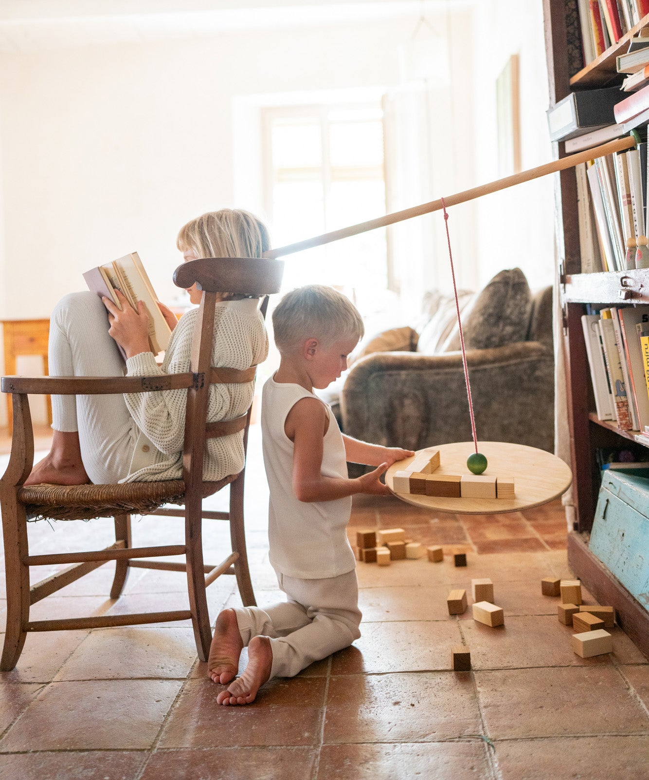 A child playing with the Grapat wooden pendulum set with wooden blocks on a terracotta floor next to a child sitting on a wooden chair with different handcrafted blocks scattered on the terracotta floor.