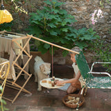 A child sitting outside with a dog playing with the Grapat wooden pendulum set balancing on a wooden clothes horse and garden chair with the different handcrafted wooden blocks balancing on the wooden board and in a woven basket on a stone floor. 