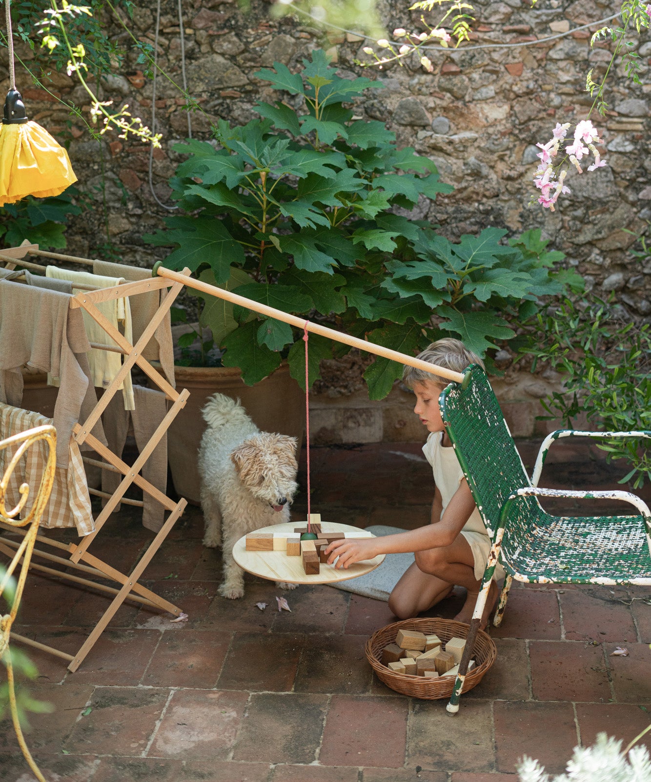A child sitting outside with a dog playing with the Grapat wooden pendulum set balancing on a wooden clothes horse and garden chair with the different handcrafted wooden blocks balancing on the wooden board and in a woven basket on a stone floor. 