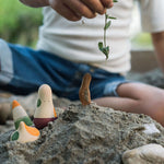 Close up of a hand holding a green plant stem above some sand with the Grapat wooden ooh lala figures sticking out