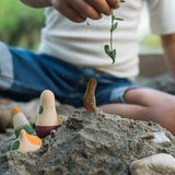 Close up of a hand holding a green plant stem above some sand with the Grapat wooden ooh lala figures sticking out
