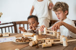 2 children sitting next to a wooden dining table stacking the different handcrafted, painted tubes and planks next to some fallen wooden tubes and planks. 