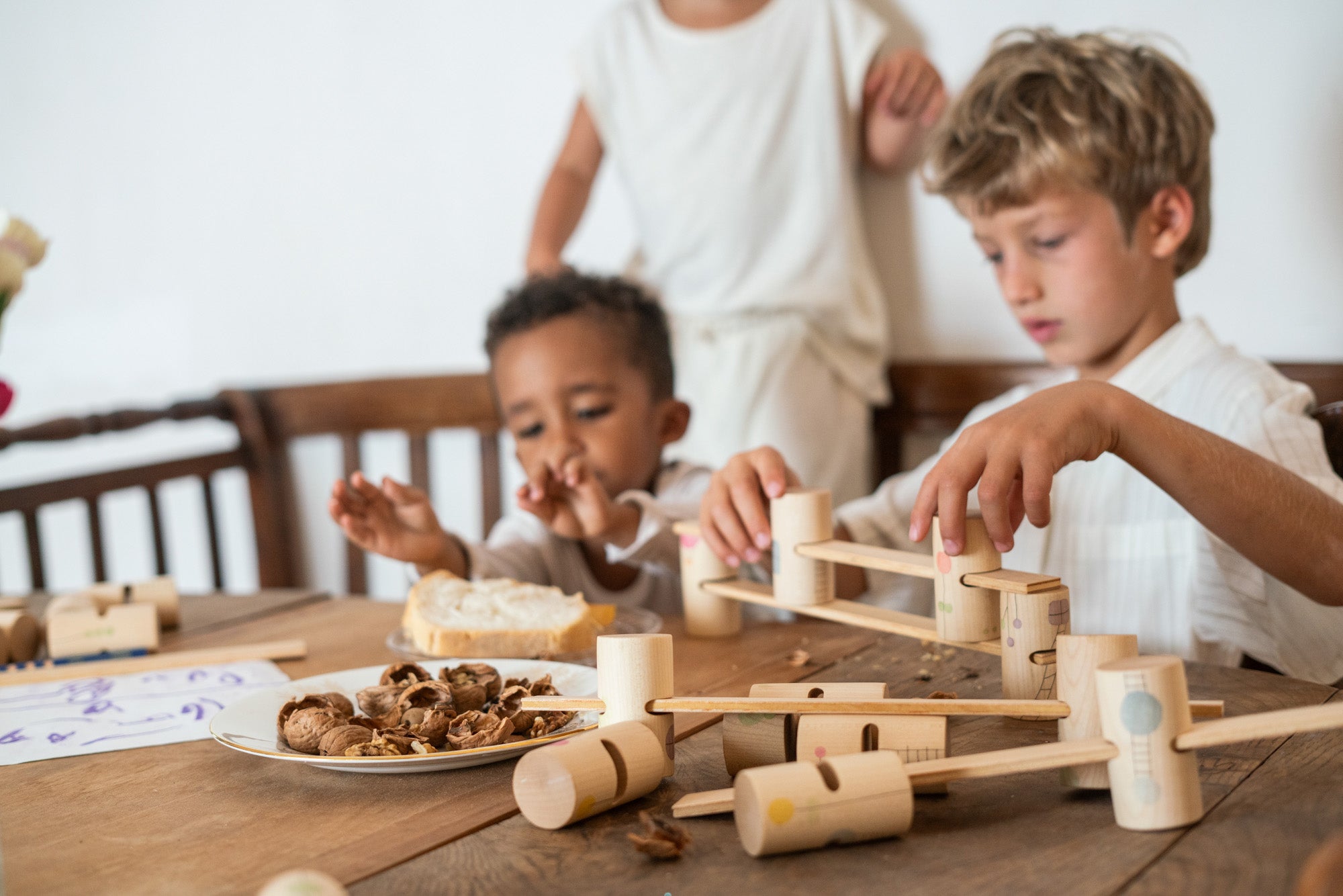 2 children sitting next to a wooden dining table stacking the different handcrafted, painted tubes and planks next to some fallen wooden tubes and planks. 
