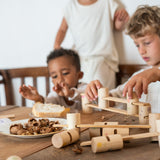 2 children sitting next to a wooden dining table stacking the different handcrafted, painted tubes and planks next to some fallen wooden tubes and planks. 
