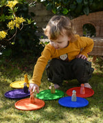 Grapat rainbow dishes placed on the grass, a child kneeling behind them placing animal crew peg dolls on the plates. 