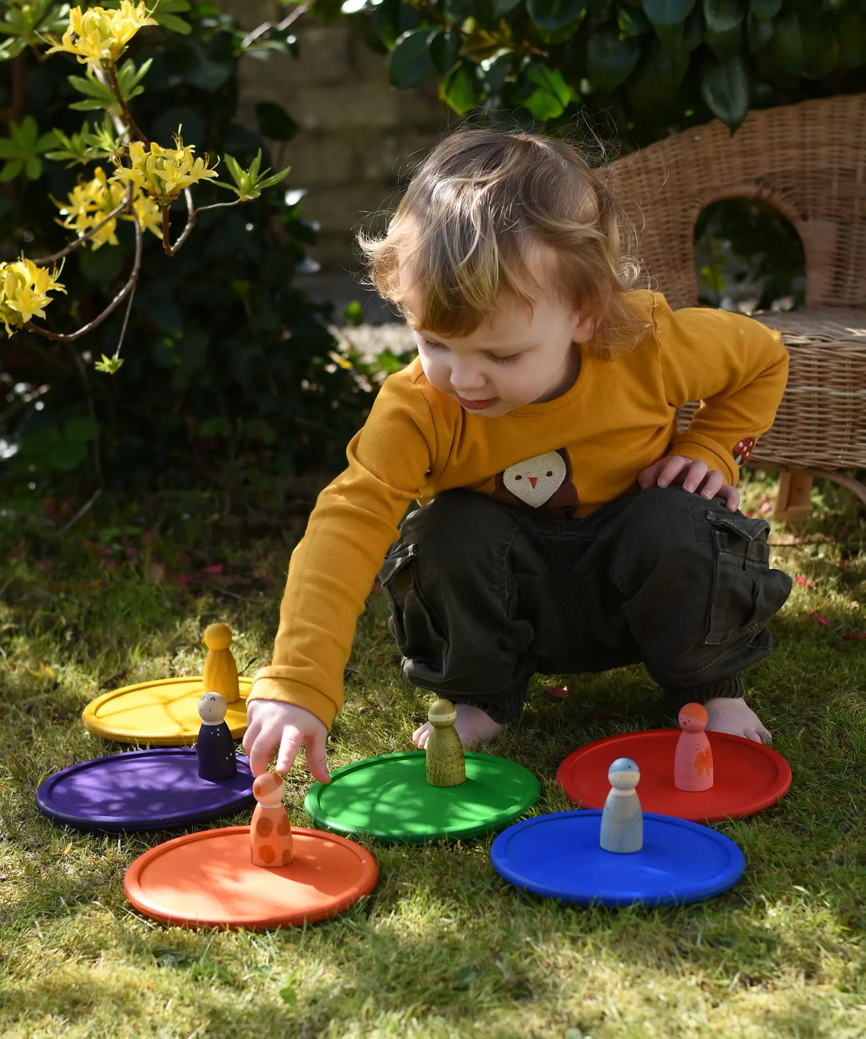 Grapat rainbow dishes placed on the grass, a child kneeling behind them placing animal crew peg dolls on the plates. 